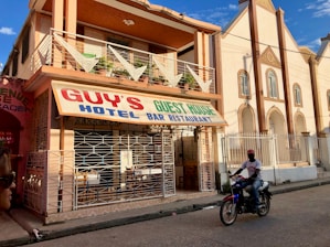 A street-side scene with a two-story building featuring a sign for Guy's Hotel, Guest House, Bar, and Restaurant. The building has a balcony with decorative triangular elements and is painted in earth tones. In front, a person rides a motorcycle on the paved road. Adjacent buildings are visible, one of which has an arched architectural design.