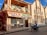 A street-side scene with a two-story building featuring a sign for Guy's Hotel, Guest House, Bar, and Restaurant. The building has a balcony with decorative triangular elements and is painted in earth tones. In front, a person rides a motorcycle on the paved road. Adjacent buildings are visible, one of which has an arched architectural design.