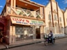A street-side scene with a two-story building featuring a sign for Guy's Hotel, Guest House, Bar, and Restaurant. The building has a balcony with decorative triangular elements and is painted in earth tones. In front, a person rides a motorcycle on the paved road. Adjacent buildings are visible, one of which has an arched architectural design.