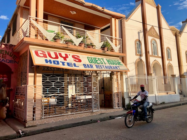 A street-side scene with a two-story building featuring a sign for Guy's Hotel, Guest House, Bar, and Restaurant. The building has a balcony with decorative triangular elements and is painted in earth tones. In front, a person rides a motorcycle on the paved road. Adjacent buildings are visible, one of which has an arched architectural design.
