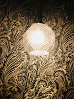 A textured spherical lamp emits a warm glow against a richly patterned wallpaper with intricate floral and paisley designs in dark tones.