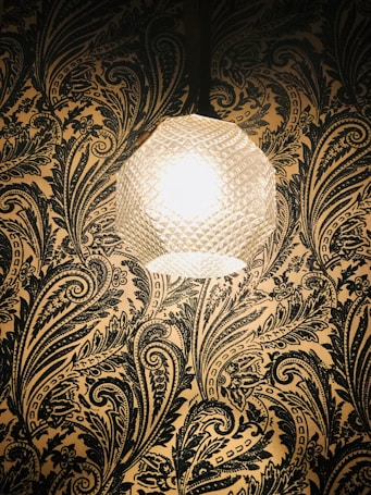 A textured spherical lamp emits a warm glow against a richly patterned wallpaper with intricate floral and paisley designs in dark tones.