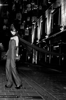 A model in an elegant long gown and gloves stands on a tiled walkway. Suspended birdcages hang above, casting shadows and providing a dramatic backdrop. The setting is urban, with modern building facades and bright, stark lighting adding to the high-fashion ambiance.