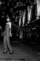 A model in an elegant long gown and gloves stands on a tiled walkway. Suspended birdcages hang above, casting shadows and providing a dramatic backdrop. The setting is urban, with modern building facades and bright, stark lighting adding to the high-fashion ambiance.