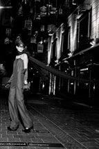 A model in an elegant long gown and gloves stands on a tiled walkway. Suspended birdcages hang above, casting shadows and providing a dramatic backdrop. The setting is urban, with modern building facades and bright, stark lighting adding to the high-fashion ambiance.