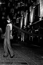 A model in an elegant long gown and gloves stands on a tiled walkway. Suspended birdcages hang above, casting shadows and providing a dramatic backdrop. The setting is urban, with modern building facades and bright, stark lighting adding to the high-fashion ambiance.