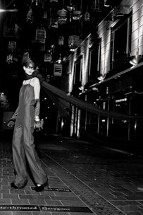 A model in an elegant long gown and gloves stands on a tiled walkway. Suspended birdcages hang above, casting shadows and providing a dramatic backdrop. The setting is urban, with modern building facades and bright, stark lighting adding to the high-fashion ambiance.