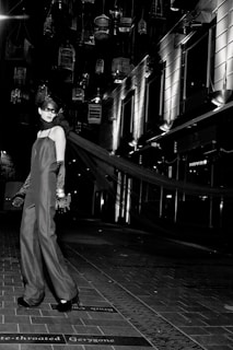A model in an elegant long gown and gloves stands on a tiled walkway. Suspended birdcages hang above, casting shadows and providing a dramatic backdrop. The setting is urban, with modern building facades and bright, stark lighting adding to the high-fashion ambiance.