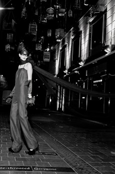 A model in an elegant long gown and gloves stands on a tiled walkway. Suspended birdcages hang above, casting shadows and providing a dramatic backdrop. The setting is urban, with modern building facades and bright, stark lighting adding to the high-fashion ambiance.