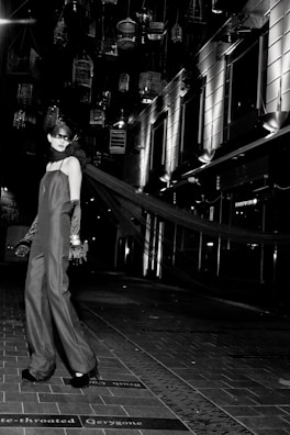 A model in an elegant long gown and gloves stands on a tiled walkway. Suspended birdcages hang above, casting shadows and providing a dramatic backdrop. The setting is urban, with modern building facades and bright, stark lighting adding to the high-fashion ambiance.