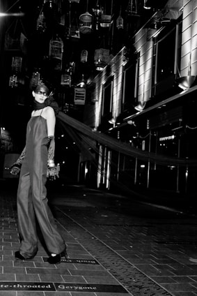A model in an elegant long gown and gloves stands on a tiled walkway. Suspended birdcages hang above, casting shadows and providing a dramatic backdrop. The setting is urban, with modern building facades and bright, stark lighting adding to the high-fashion ambiance.