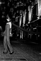 A model in an elegant long gown and gloves stands on a tiled walkway. Suspended birdcages hang above, casting shadows and providing a dramatic backdrop. The setting is urban, with modern building facades and bright, stark lighting adding to the high-fashion ambiance.