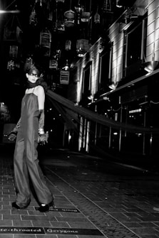 A model in an elegant long gown and gloves stands on a tiled walkway. Suspended birdcages hang above, casting shadows and providing a dramatic backdrop. The setting is urban, with modern building facades and bright, stark lighting adding to the high-fashion ambiance.