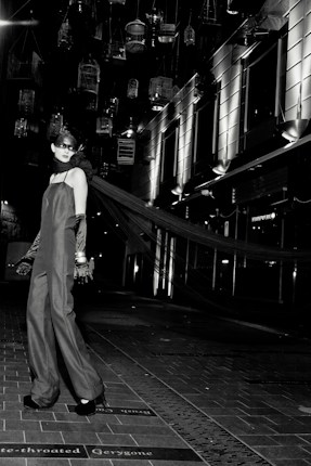 A model in an elegant long gown and gloves stands on a tiled walkway. Suspended birdcages hang above, casting shadows and providing a dramatic backdrop. The setting is urban, with modern building facades and bright, stark lighting adding to the high-fashion ambiance.