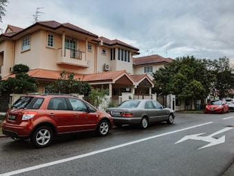 A residential street with a two-story house featuring a terracotta-tiled roof and cream-colored walls. Three cars are parked along the street, with a red SUV in the foreground and two sedans behind it. Lush, green trees partially obscure the view of the house's front yard.