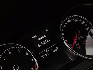 A close-up of a car dashboard displaying various gauges, including a speedometer and tachometer. The digital display reads a range of 520 km and shows time, temperature, and trip distance information. The gauges have white numbers and red needles on black backgrounds.