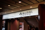 An illuminated store sign featuring both Chinese characters and English text. The ceiling above has recessed lights, and there are two people underneath the sign.