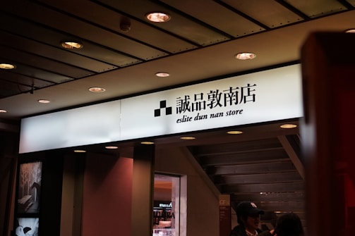 An illuminated store sign featuring both Chinese characters and English text. The ceiling above has recessed lights, and there are two people underneath the sign.