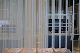 A modern interior space with sheer white curtains partially concealing a large flat-screen monitor and some shelving. In the foreground, newspapers with colorful prints are visible on a smooth table surface. The setting appears minimalistic and clean.