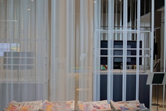 A modern interior space with sheer white curtains partially concealing a large flat-screen monitor and some shelving. In the foreground, newspapers with colorful prints are visible on a smooth table surface. The setting appears minimalistic and clean.