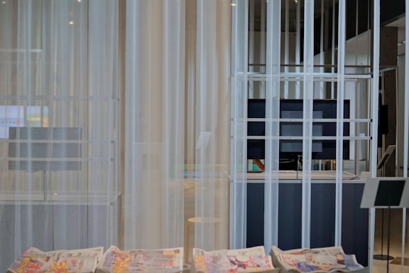 A modern interior space with sheer white curtains partially concealing a large flat-screen monitor and some shelving. In the foreground, newspapers with colorful prints are visible on a smooth table surface. The setting appears minimalistic and clean.