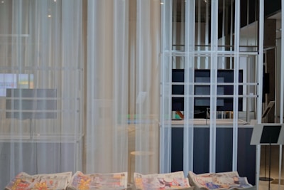 A modern interior space with sheer white curtains partially concealing a large flat-screen monitor and some shelving. In the foreground, newspapers with colorful prints are visible on a smooth table surface. The setting appears minimalistic and clean.
