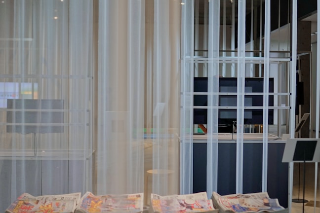 A modern interior space with sheer white curtains partially concealing a large flat-screen monitor and some shelving. In the foreground, newspapers with colorful prints are visible on a smooth table surface. The setting appears minimalistic and clean.