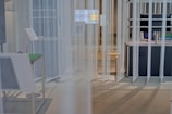 The image depicts a modern, minimalist space with vertical translucent panels hanging from the ceiling. A worktable is present on the right, accompanied by a chair, and there's a visible screen on the desk. A person is partially visible behind the desk, possibly working on a computer.
