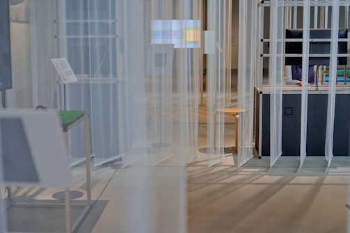 The image depicts a modern, minimalist space with vertical translucent panels hanging from the ceiling. A worktable is present on the right, accompanied by a chair, and there's a visible screen on the desk. A person is partially visible behind the desk, possibly working on a computer.