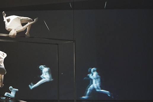 A surreal scene featuring a geometric, angular sculpture of a human figure positioned upside-down near the top left. In the background, a series of semi-transparent, dynamic images depict a human figure in various stages of running, conveying motion. The setting is mostly dark, emphasizing the light shapes and movement.