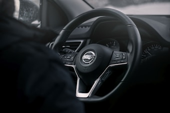 A close-up view of a Nissan car steering wheel, featuring the brand logo prominently in the center. The dashboard displays several dials and buttons. The image is mostly dark, creating a sleek and modern feel.
