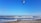 Kitesurfers navigate the waves along a sandy beach under a clear blue sky. Colorful kites sail above the ocean, capturing the lively energy of the sport. Pebbles scatter across the shoreline, adding texture to the scene.