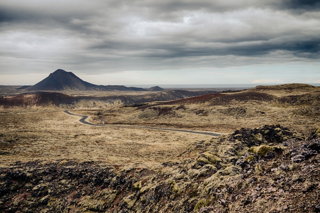 A vast, barren landscape with a winding road cutting through it. Rugged terrain is covered in moss and volcanic rocks. In the background, a prominent mountain rises under a cloudy sky, creating a dramatic horizon.