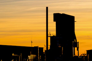 Silhouettes of industrial machinery fading into colorful burnt orange and golden yellow tie-dye clouds at sunset.