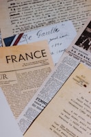 A collection of vintage newspapers and handwritten letters, mostly written in French, are laid out overlapping each other. The papers exhibit different fonts and styles, reflecting historical and artistic significance.