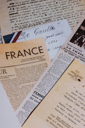 A collection of vintage newspapers and handwritten letters, mostly written in French, are laid out overlapping each other. The papers exhibit different fonts and styles, reflecting historical and artistic significance.