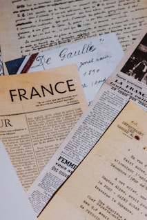 A collection of vintage newspapers and handwritten letters, mostly written in French, are laid out overlapping each other. The papers exhibit different fonts and styles, reflecting historical and artistic significance.