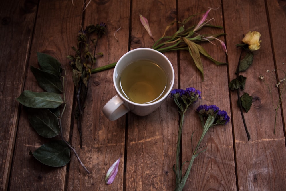 Tasse en céramique blanche avec du thé sur une surface en bois à côté de plantes à feuilles vertes et de fleurs à pétales