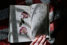 An open botanical book sits on a textured fabric surface, displaying illustrations and descriptions of various plants. Pressed flowers lie on one of the pages, while soft natural light illuminates the scene. A person's hand gently holds the corner of the book.