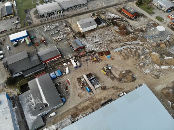 An aerial view of an industrial area with multiple warehouses and buildings, surrounded by dirt roads and construction materials. Snow is partially covering the ground and rooftops. Several trucks and machinery are parked around the site.