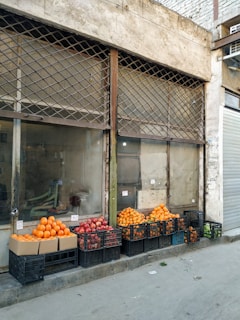 Stacks of fresh fruits, including oranges and pomegranates, are displayed in black crates and cardboard boxes outside a run-down building with barred windows. The setup is on a concrete sidewalk with a closed metal roller shutter on one side.