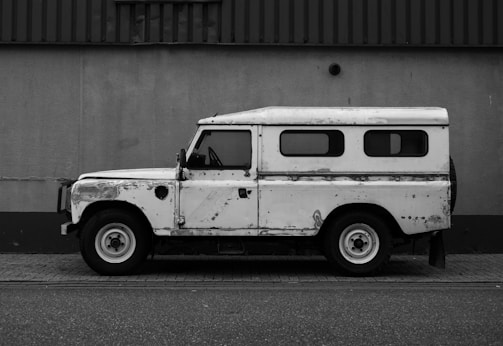 A vintage Land Rover vehicle parked against a plain wall, exhibiting signs of wear and rust. The vehicle has a boxy shape with a white body, visible windows along the side, and noticeable wear on the exterior paint.