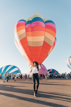 A vibrant hot air balloon festival with multiple colorful balloons in the background. A woman in a white blouse and dark jeans walks confidently, wearing a wide-brimmed hat. A crowd of people observes the preparation and launch of the balloons.