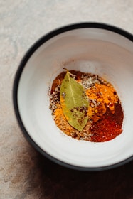 An overhead view of a white bowl containing a variety of colorful spices, with a bay leaf placed on top. The spices are neatly arranged in separate sections within the bowl.