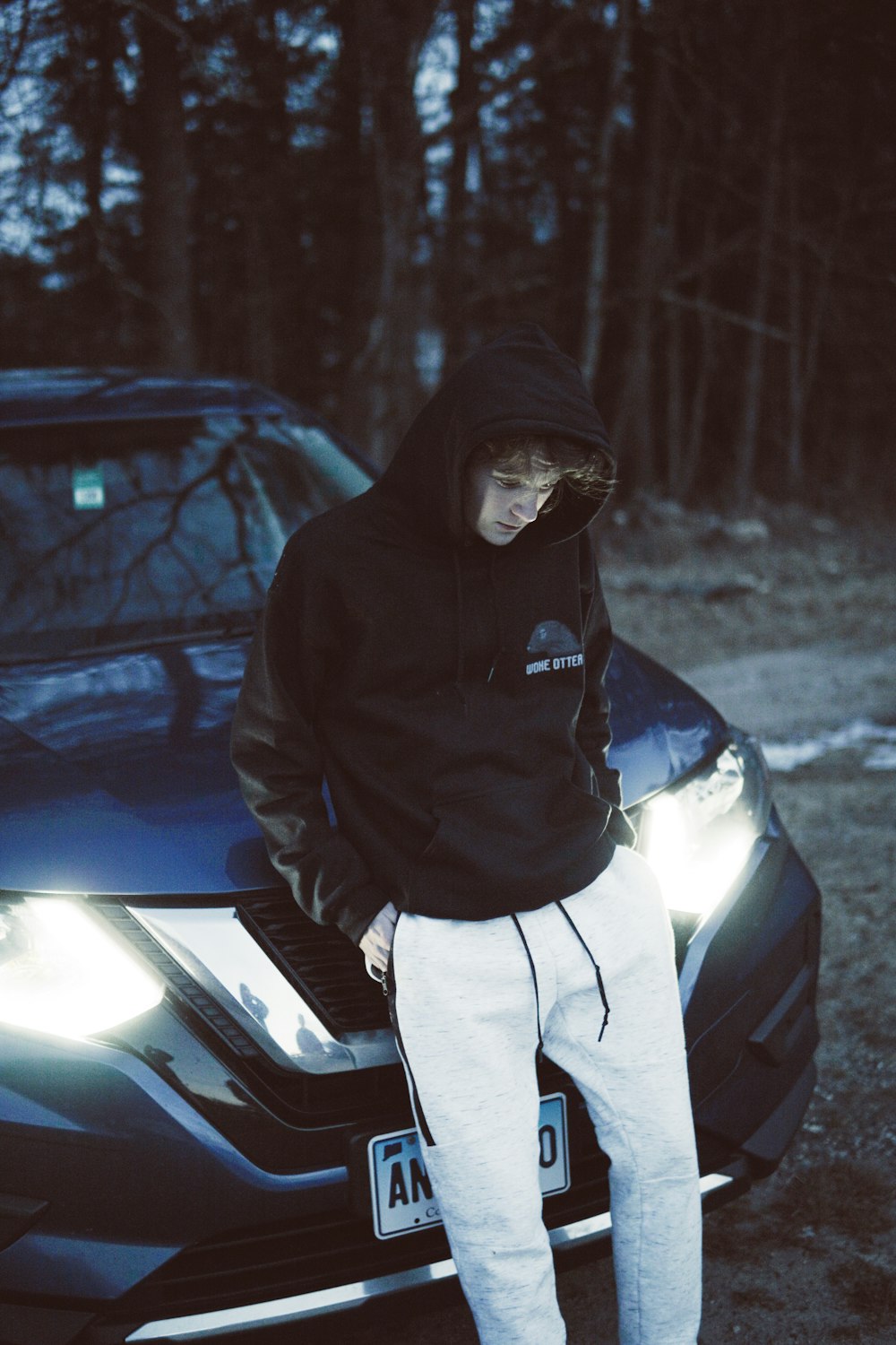 man wearing black hoodie leaning on blue car near trees