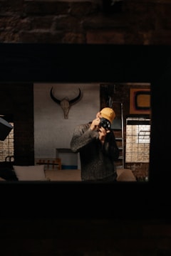 A person wearing a grey sweater and yellow cap holds a camera, capturing a photograph. The room features exposed brick walls, a mounted animal skull with horns, and a window with a grid frame. A sofa with cushions is partially visible. The setting conveys a rustic and artistic ambiance.