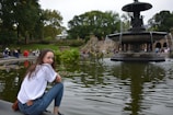 A young person sits by a serene fountain in a park. The fountain has multiple tiers with water cascading down, surrounded by lush greenery. In the background, a variety of people are leisurely enjoying the park, some walking up stone steps.