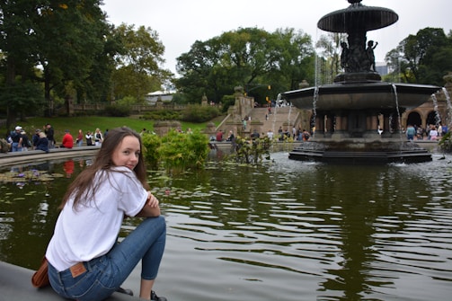 A young person sits by a serene fountain in a park. The fountain has multiple tiers with water cascading down, surrounded by lush greenery. In the background, a variety of people are leisurely enjoying the park, some walking up stone steps.
