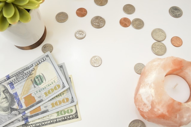 A collection of US currency is spread out on a white surface, including several hundred-dollar bills and a variety of coins. A small potted succulent plant is positioned on the left side, while a pink Himalayan salt candle holder is on the right.