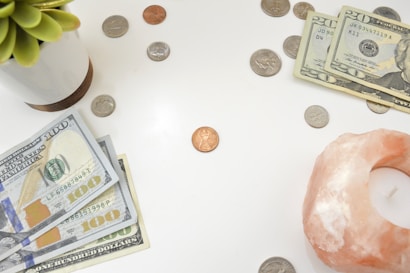 A white surface is scattered with US currency, including dollar bills and coins. A stack of one hundred and twenty dollar bills is visible alongside various coins, including quarters and pennies. On the left edge, a small succulent in a white pot is present, and on the right, a pink himalayan salt candle holder holds a small candle.